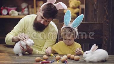 父子俩正在<strong>画</strong>复活节<strong>彩蛋</strong>.. 可爱的小男孩戴着兔子耳朵。 木制背景的复活节<strong>彩蛋</strong>。 男人